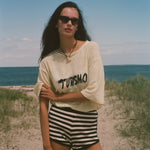 Woman standing on a beach wearing sunglasses and a yellow shirt with 'TURISMO' embroidered across the front.