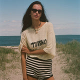 Woman standing on a beach wearing sunglasses and a yellow shirt with 'TURISMO' embroidered across the front.