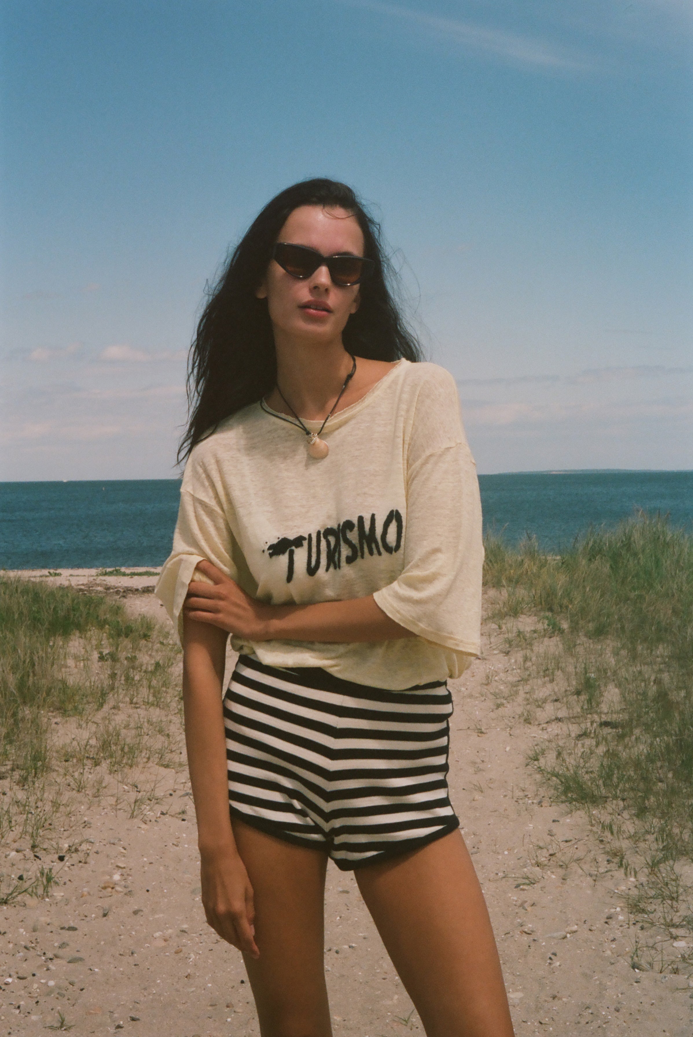 Woman standing on a beach wearing sunglasses and a yellow shirt with 'TURISMO' embroidered across the front.