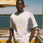 A man wearing a white graphic t-shirt with a blue and yellow fish design and yellow shorts, standing on a sunny boat deck with water and a blue sky in the background.