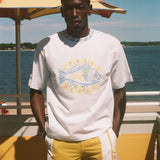A man standing outdoors by the water wearing a white graphic T-shirt with a large fish design and yellow shorts, with a beach umbrella and blue sky in the background.