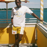 A man stands on a sunny outdoor deck by the water wearing a white graphic T-shirt and yellow shorts with white side stripes. An orange-and-white striped umbrella, yellow railings, and blue water are visible in the background.