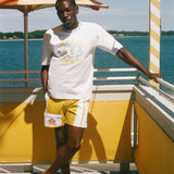 A man stands on a sunny outdoor deck by the water wearing a white graphic T-shirt and yellow shorts with white side stripes. An orange-and-white striped umbrella, yellow railings, and blue water are visible in the background.