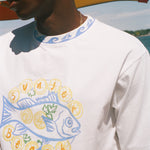 A close-up of a person wearing a white graphic t-shirt with a blue and yellow 'Sunset Beach' fish design, set against a sunny outdoor background with a beach umbrella.