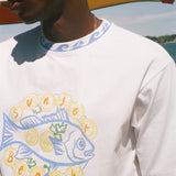A close-up of a person wearing a white graphic t-shirt with a blue and yellow 'Sunset Beach' fish design, set against a sunny outdoor background with a beach umbrella.