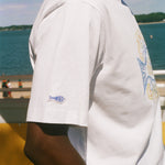 A close-up side view of a person wearing a white short-sleeve t-shirt with a blue and yellow graphic on the back and a small 'Tombolo' logo on the sleeve, set against a background of water and a yellow structure.