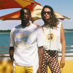 Two models standing outdoors by the water under a red and yellow striped beach umbrella. The model on the left wears a white graphic t-shirt with a blue fish and yellow text, yellow shorts, and a white pocket.