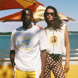 Two models standing outdoors by the water under a red and yellow striped beach umbrella. The model on the left wears a white graphic t-shirt with a blue fish and yellow text, yellow shorts, and a white pocket.