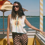 A woman wearing a sleeveless white top with a yellow sun graphic and brown polka-dot pants stands on a sunny outdoor deck with water and umbrellas in the background.