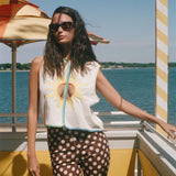 A woman wearing a sleeveless white top with a yellow sun graphic and brown polka-dot pants stands on a sunny outdoor deck with water and umbrellas in the background.