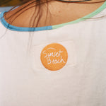 Close-up of the back of a white shirt worn by a person, featuring a circular orange patch with 'Sunset Beach' embroidered in white script. 