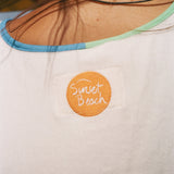 Close-up of the back of a white shirt worn by a person, featuring a circular orange patch with 'Sunset Beach' embroidered in white script. 