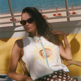 A woman wearing a white sleeveless top with a yellow sun graphic and brown polka-dot pants, seated in a wicker chair with a beach and ocean in the background.