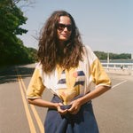 A woman with long, windblown brown hair and dark sunglasses stands on an outdoor road wearing a yellow and white striped shirt.