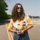 A woman with long, windblown brown hair and dark sunglasses stands on an outdoor road wearing a yellow and white striped shirt.