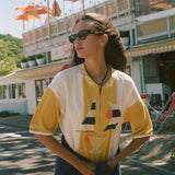 A woman wearing a yellow and white color-blocked short-sleeve shirt with a sailboat and cocktail graphic, styled with dark sunglasses and a navy blue jacket tied around her waist, standing outdoors in front of a building with orange umbrellas.