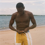 A shirtless man standing on a sandy beach, looking down while adjusting yellow and white swim trunks with a drawstring. The background shows the ocean and a pale sky.
