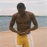 A shirtless man standing on a sandy beach, looking down while adjusting yellow and white swim trunks with a drawstring. The background shows the ocean and a pale sky.