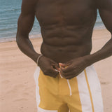 A shirtless man standing on a sandy beach wearing yellow and white swim trunks, with the ocean and sky in the background.