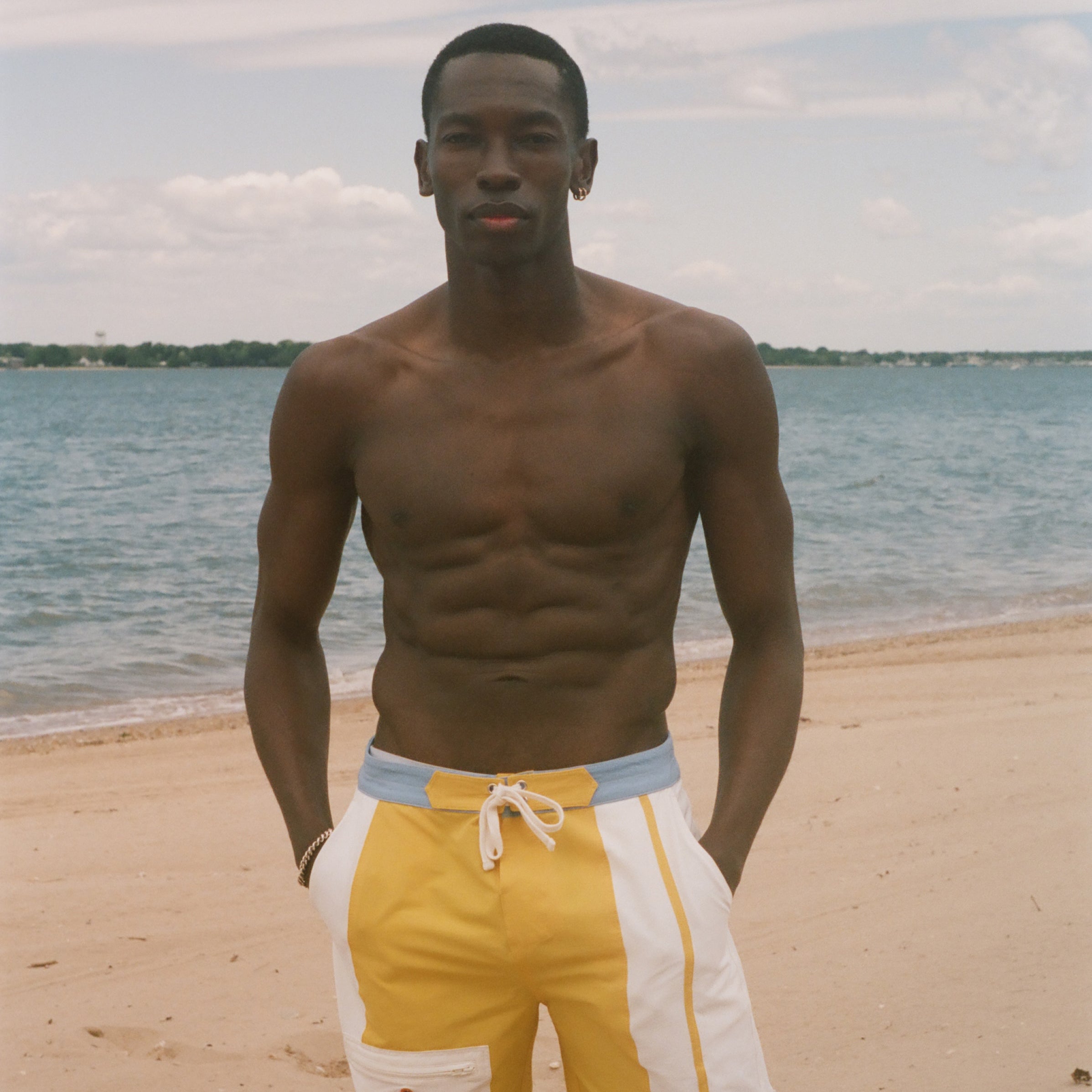 A shirtless man standing on a sandy beach wearing yellow and white swim trunks with a light blue waistband, with the ocean and sky behind him.