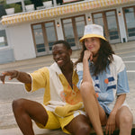 Two models sitting on wooden planks in front of a beachfront restaurant, wearing colorful summer outfits. One model wears a yellow and white short-sleeve shirt and matching shorts, while the other wears a white, blue, and orange graphic t-shirt with a yellow bucket hat.