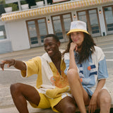 Two models sitting on wooden planks in front of a beachfront restaurant, wearing colorful summer outfits. One model wears a yellow and white short-sleeve shirt and matching shorts, while the other wears a white, blue, and orange graphic t-shirt with a yellow bucket hat.