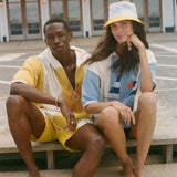 Two models sitting on concrete steps outdoors, wearing coordinated beach-style outfits. The person on the left wears a yellow and white short-sleeve top and matching shorts.