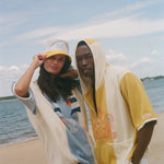 Two models standing on a sandy beach wearing matching yellow and off-white hooded beach poncho-style tops under a blue sky.