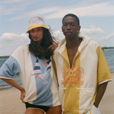 Two models posing on a sandy beach wearing matching color-blocked hoodies and shorts sets. The set features a color-blocked design of cream, yellow, and light blue, with a tropical cocktail graphic on the woman's hoodie and a stylized graphic on the man's hoodie.