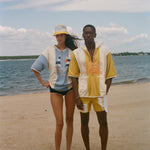 Two models standing on a sandy beach wearing matching two-piece swimwear sets. The woman wears a light blue and white set with a yellow and white cap, and the man wears a yellow and white set with a yellow and white cap.