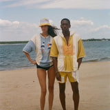 Two models standing on a sandy beach wearing matching two-piece swimwear sets. The woman wears a light blue and white set with a yellow and white cap, and the man wears a yellow and white set with a yellow and white cap.