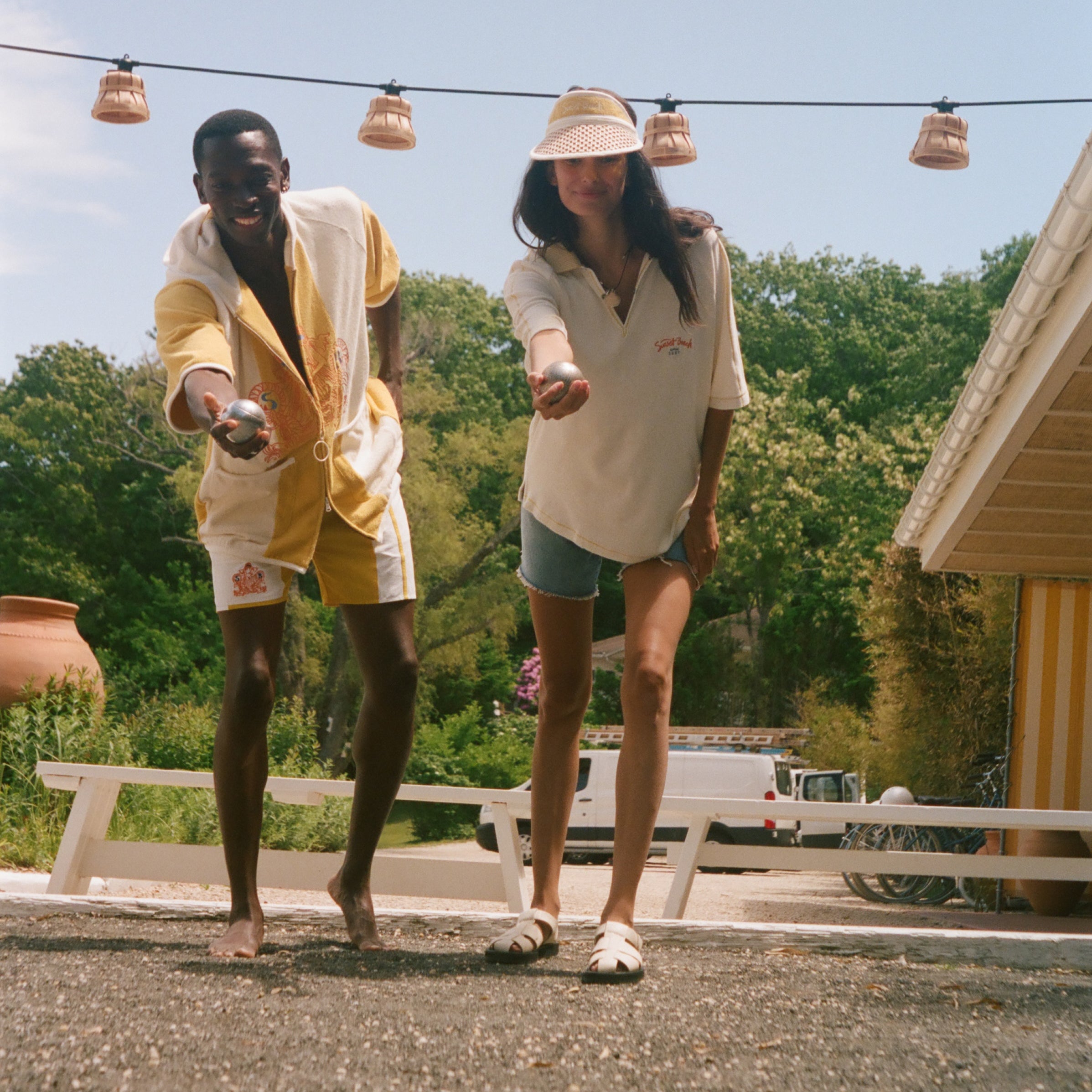 Two people outdoors on a gravel surface under a blue sky, wearing matching cream and yellow lounge sets while playing with small balls. A house, trees, and hanging string lights are visible in the background.