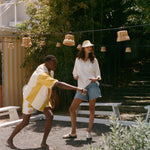 A man and a woman outdoors in a sunny garden or patio setting. The man wears a white and yellow striped shirt and shorts, while the woman wears a white long-sleeve top, denim shorts, and a straw hat.