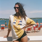 A woman stands on a beach wearing an oversized yellow and white graphic t-shirt, black swim trunks, and sunglasses, with the ocean and lounge chairs in the background.