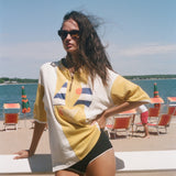 A woman stands on a beach wearing an oversized yellow and white graphic t-shirt, black swim trunks, and sunglasses, with the ocean and lounge chairs in the background.