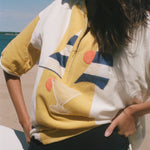 A woman at the beach wearing a yellow and white short-sleeve zip-up shirt with a graphic print of a cocktail glass and blue geometric shapes, paired with black swim shorts.