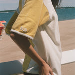 A woman stands on a beach wearing an oversized yellow and white graphic t-shirt, black swim trunks, and sunglasses, with the ocean and lounge chairs in the background.