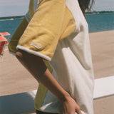 A woman stands on a beach wearing an oversized yellow and white graphic t-shirt, black swim trunks, and sunglasses, with the ocean and lounge chairs in the background.