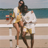 A couple posing in matching yellow and white swimwear and loungewear sets on a beach boardwalk with a white fence, ocean, and sky in the background.