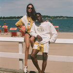 Two models posing on a beach boardwalk in front of a white railing, with the ocean, sand, and beach chairs in the background. The woman wears a yellow and white striped poncho.