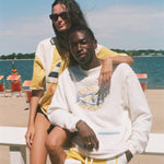 Two models posing on a white beach chair at the beach, wearing yellow and white swimwear and a white sweatshirt with a graphic print.