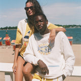 Two models posing on a white beach chair at the beach, wearing yellow and white swimwear and a white sweatshirt with a graphic print.