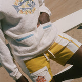 A person at a beach wearing a white terry sweatshirt with a blue and yellow fish graphic on the chest, white shorts with yellow side panels and a white front pocket featuring a small orange graphic, and dark swim trunks.