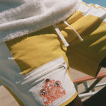 Close-up of a person wearing yellow and white board shorts with a white fleece-lined pocket featuring a red graphic print, under a white towel, with a blue sky background.
