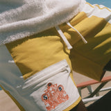 Close-up of a person wearing yellow and white board shorts with a white fleece-lined pocket featuring a red graphic print, under a white towel, with a blue sky background.