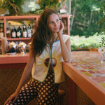 A woman posing in a vibrant, tropical-themed bar or cafe, wearing a yellow sun graphic vest over a white top and black pants with white polka dots.