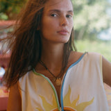 A woman wearing a sleeveless white top with a yellow sun graphic and colorful trim, photographed outdoors with a blurred green background.