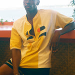 A man wearing a yellow and white short-sleeve button-up shirt with a geometric graphic on the front, paired with dark shorts, leaning against a wall in an outdoor setting.