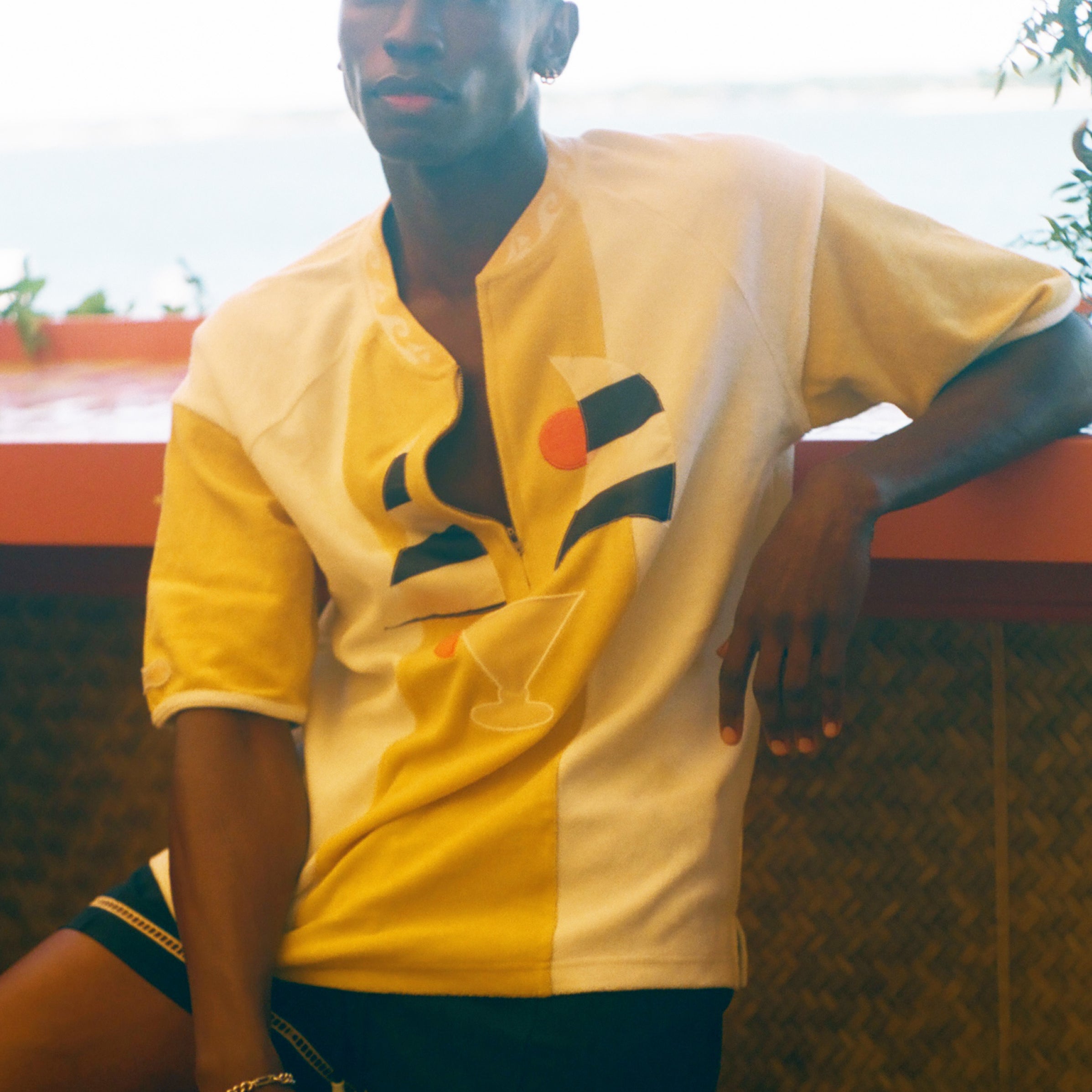A man wearing a yellow and white short-sleeve button-up shirt with a geometric graphic on the front, paired with dark shorts, leaning against a wall in an outdoor setting.