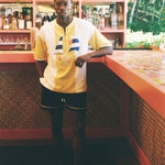 A man wearing a yellow and white short-sleeve athletic jersey with blue and red accents, paired with black athletic shorts, standing in a tropical bar environment.